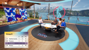 View of the virtual set created by Lightwell for BBC Sports coverage of the UEFA Womens Euros 2025. Gaby Logan sits in the presenters chair, across the desk from 3 guests. The space in which they sit is virtual, but creted to look like a modern, timber pavilion. Through the windows, we can see Lake Lucerne and mountains. This swiss landscape is also virtual.