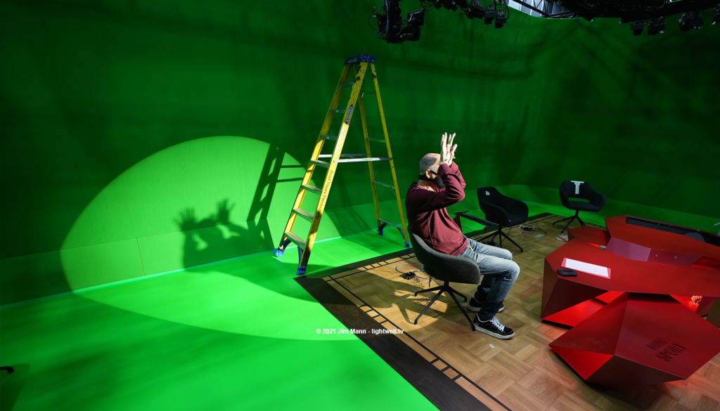 Lighting director Dave Gibson sits in a presenters chair whilst lights are focused on him during installation. In a playful interlude, he creates shadow monsters on the back wall by placing his hands besides his head.