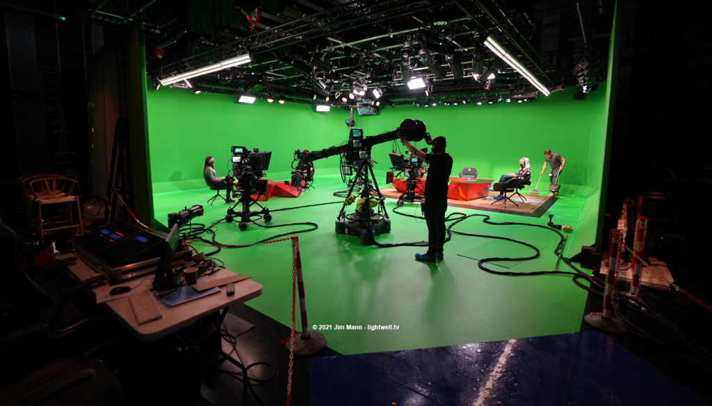 A wide view of the greenscreen studio seen from just outside the entrance. A cameraman operates the jib, whilst stand-ins sit in the presenters chairs whilst camera moves are rehearsed.
